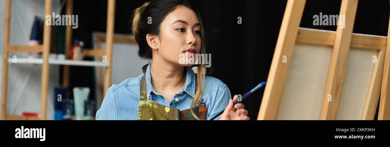 Une jeune artiste asiatique, vêtue d’un tablier, se tient dans son atelier, contemplant avec soin son prochain coup de pinceau sur une toile. Banque D'Images
