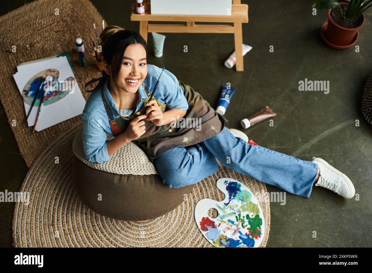 Une jeune femme asiatique, vêtue d'un tablier, s'assoit confortablement sur une chaise de sac de haricots dans son atelier d'art, tenant un pinceau et souriant. Banque D'Images