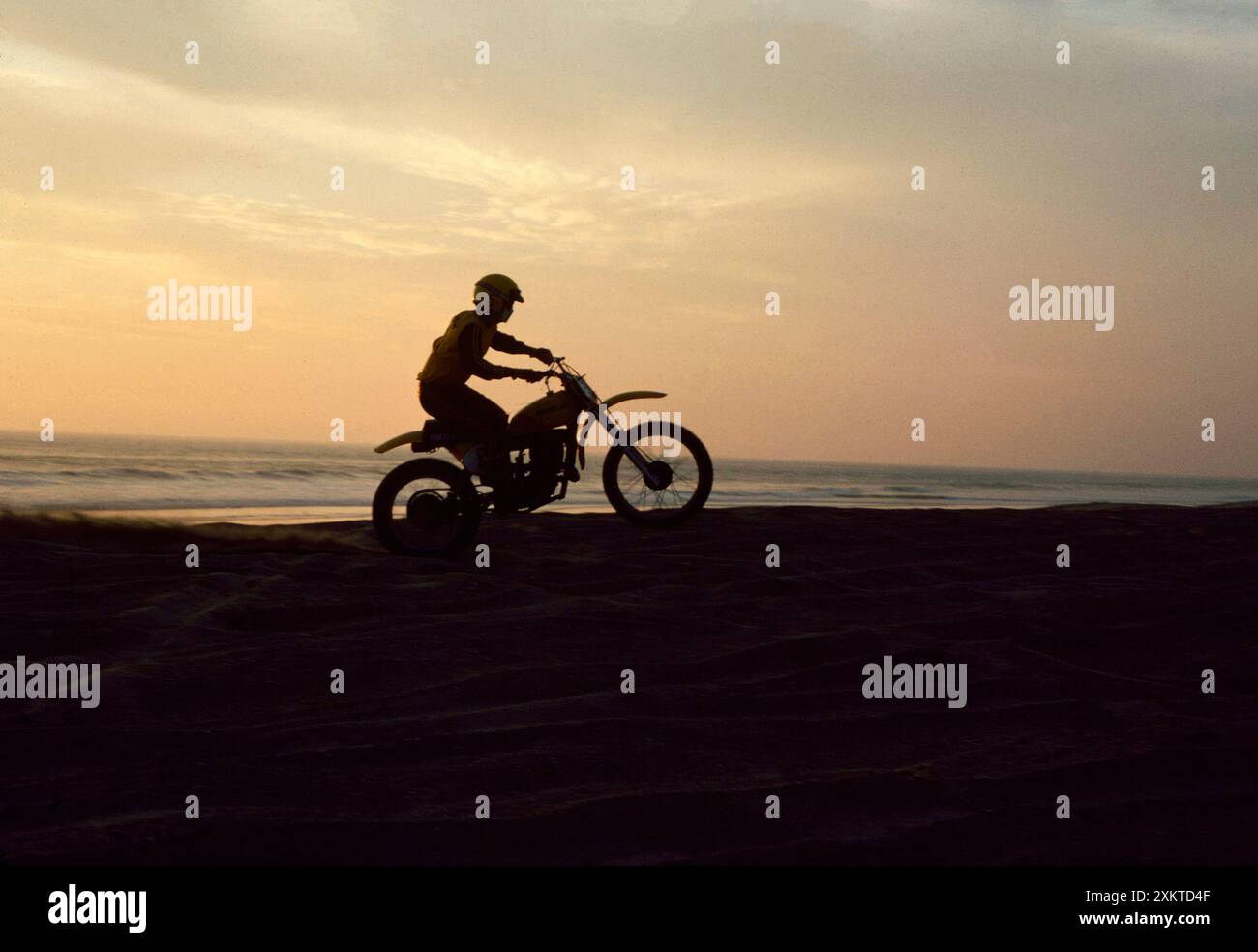 1974 Southern California Beach motocross rider Wheelie à travers le sable en silhouette au coucher du soleil Banque D'Images
