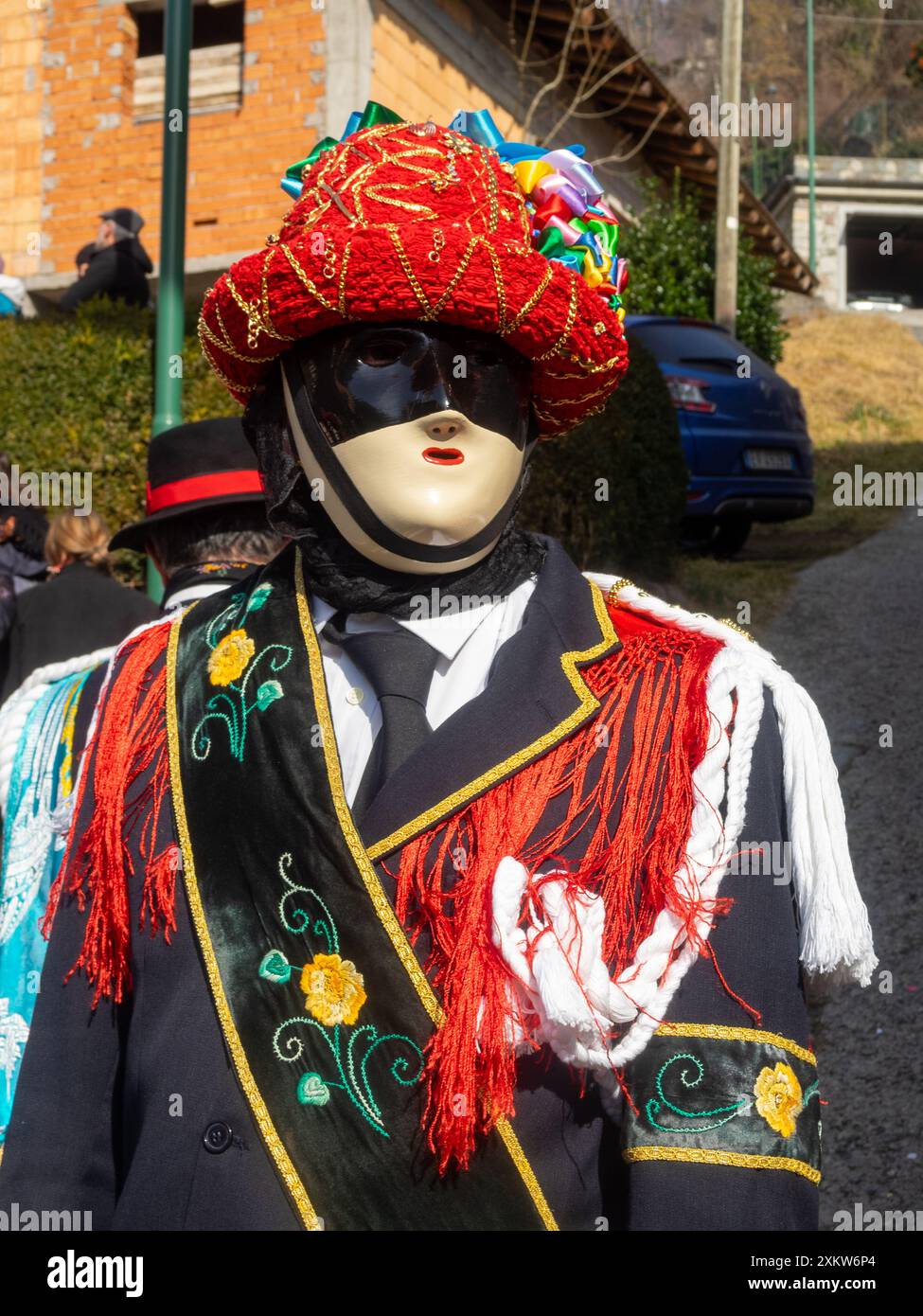Portrait de Bagolino Carnaval Balari, visage couvert par le masque ivoire et noir sans expression, tête sous un chapeau de feutre, recouvert de ruban rouge, bijoux en or et rubans colorés, portant une robe noire avec une bande de soie avec broderie de fleurs Banque D'Images