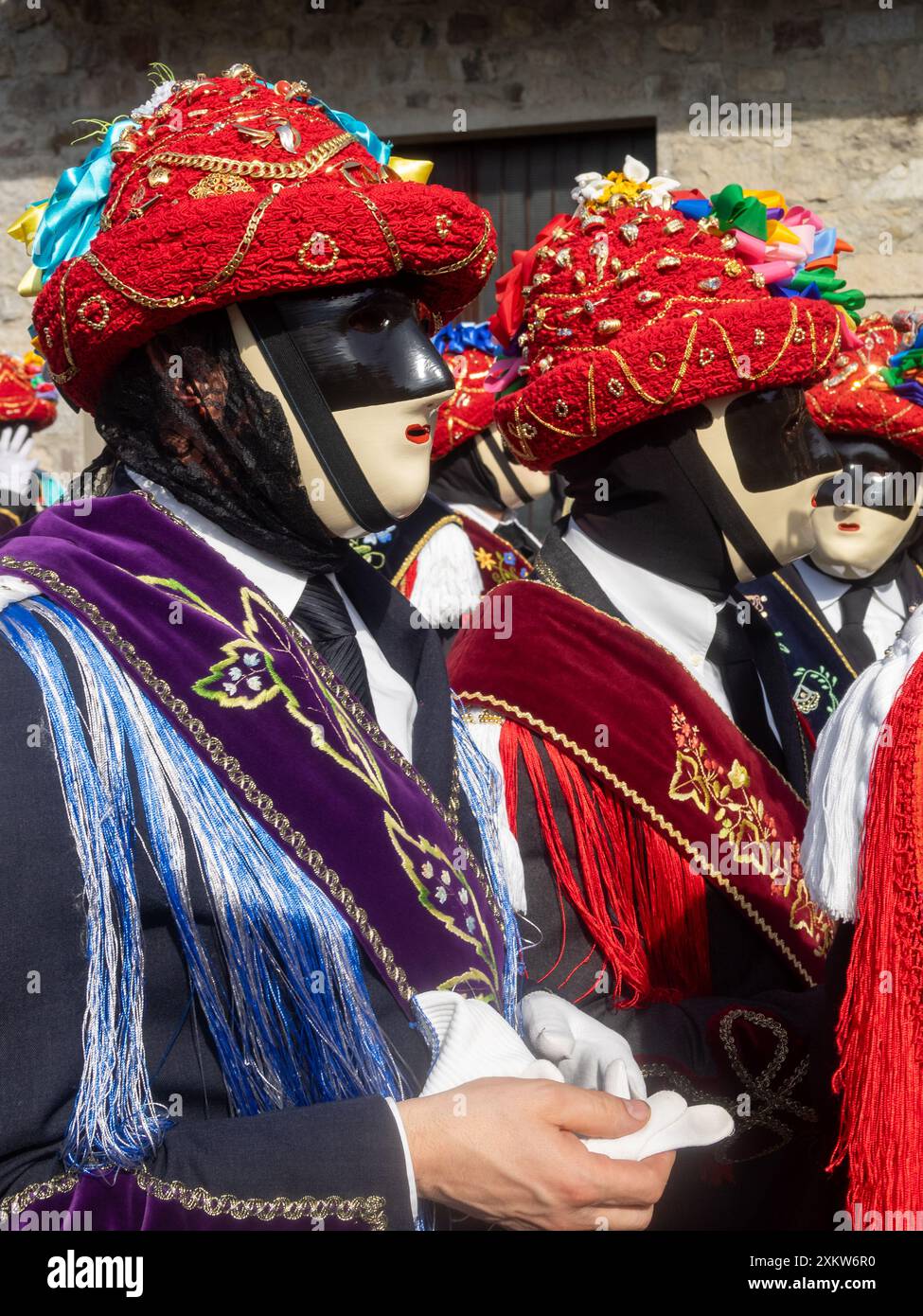 Portrait de Bagolino Carnaval Balari, visage couvert par le masque ivoire et noir sans expression, tête sous un chapeau de feutre, recouvert de ruban rouge, bijoux en or et rubans colorés, portant une robe noire avec une bande de soie avec des fleurs brodées et des gants blancs Banque D'Images