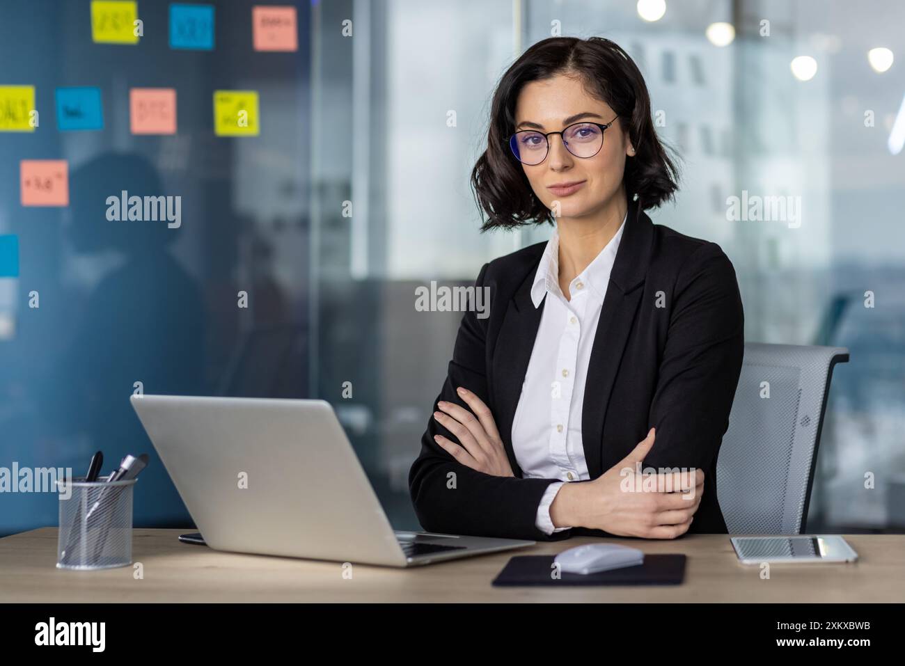 Femme d'affaires confiante avec des lunettes travaillant sur ordinateur portable dans un cadre de bureau moderne. Espace de travail professionnel avec notes adhésives et accessoires de bureau. Idéal pour les affaires, le bureau et les thèmes professionnels. Banque D'Images