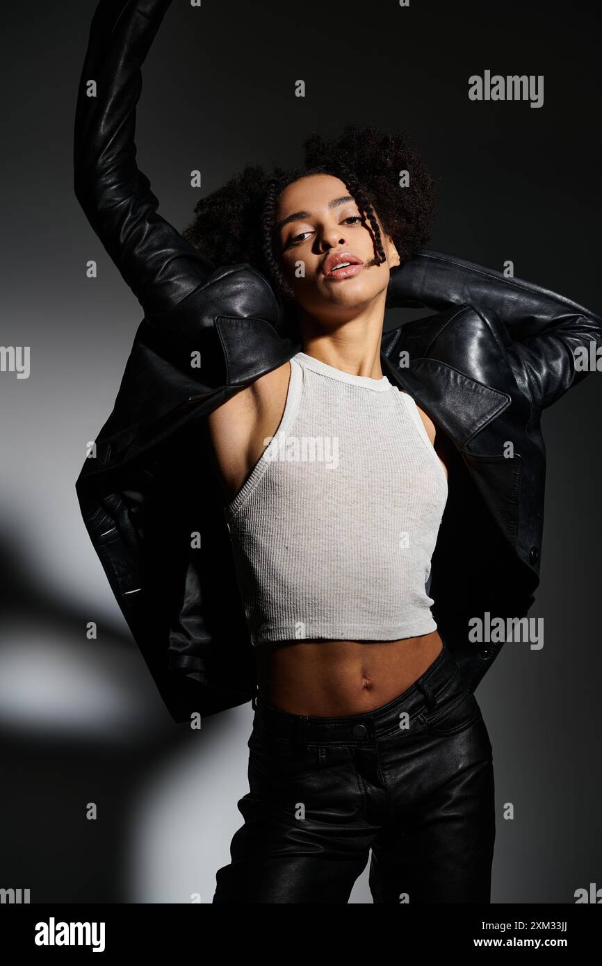 Une jeune femme afro-américaine élégante pose dans un débardeur blanc et une veste en cuir noir sur un fond gris. Banque D'Images