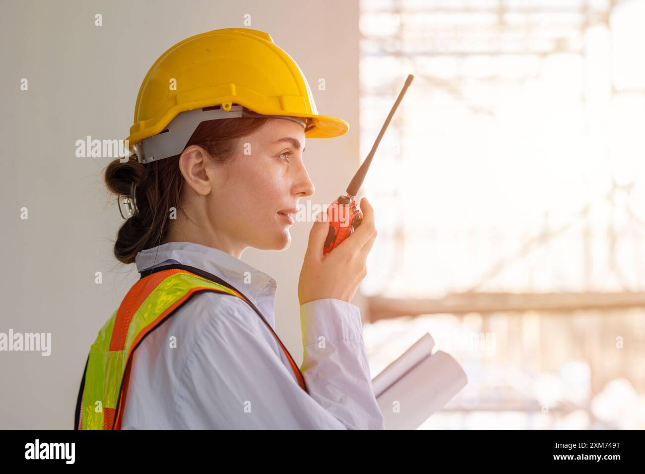 Travailleuse de la construction portant un équipement de sécurité et un casque de sécurité, communiquant à l'aide d'une radio et tenant des plans sur un chantier de construction. Banque D'Images