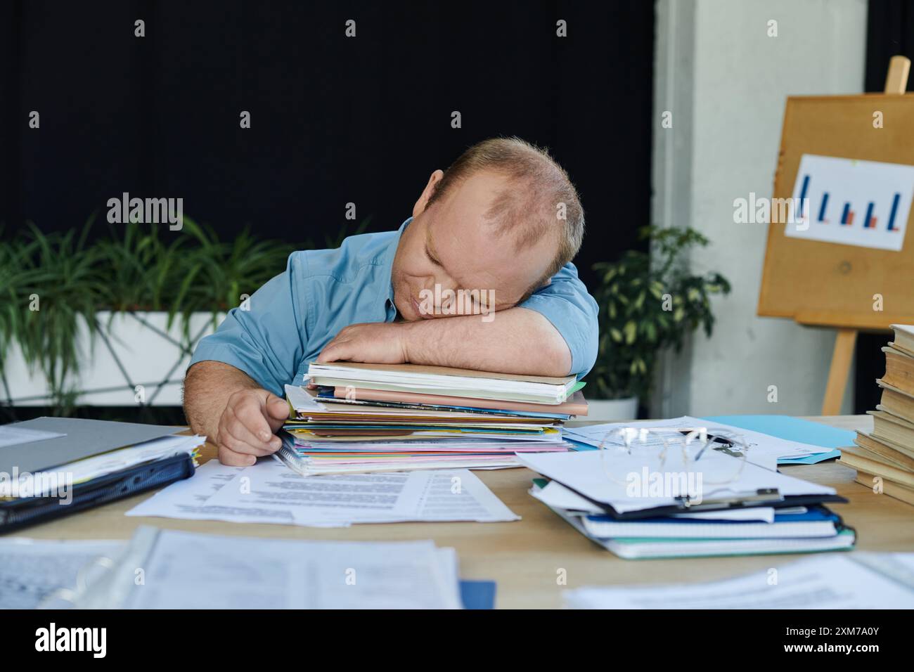 Un homme inclusif repose sa tête sur une pile de papiers tout en travaillant à son bureau. Banque D'Images