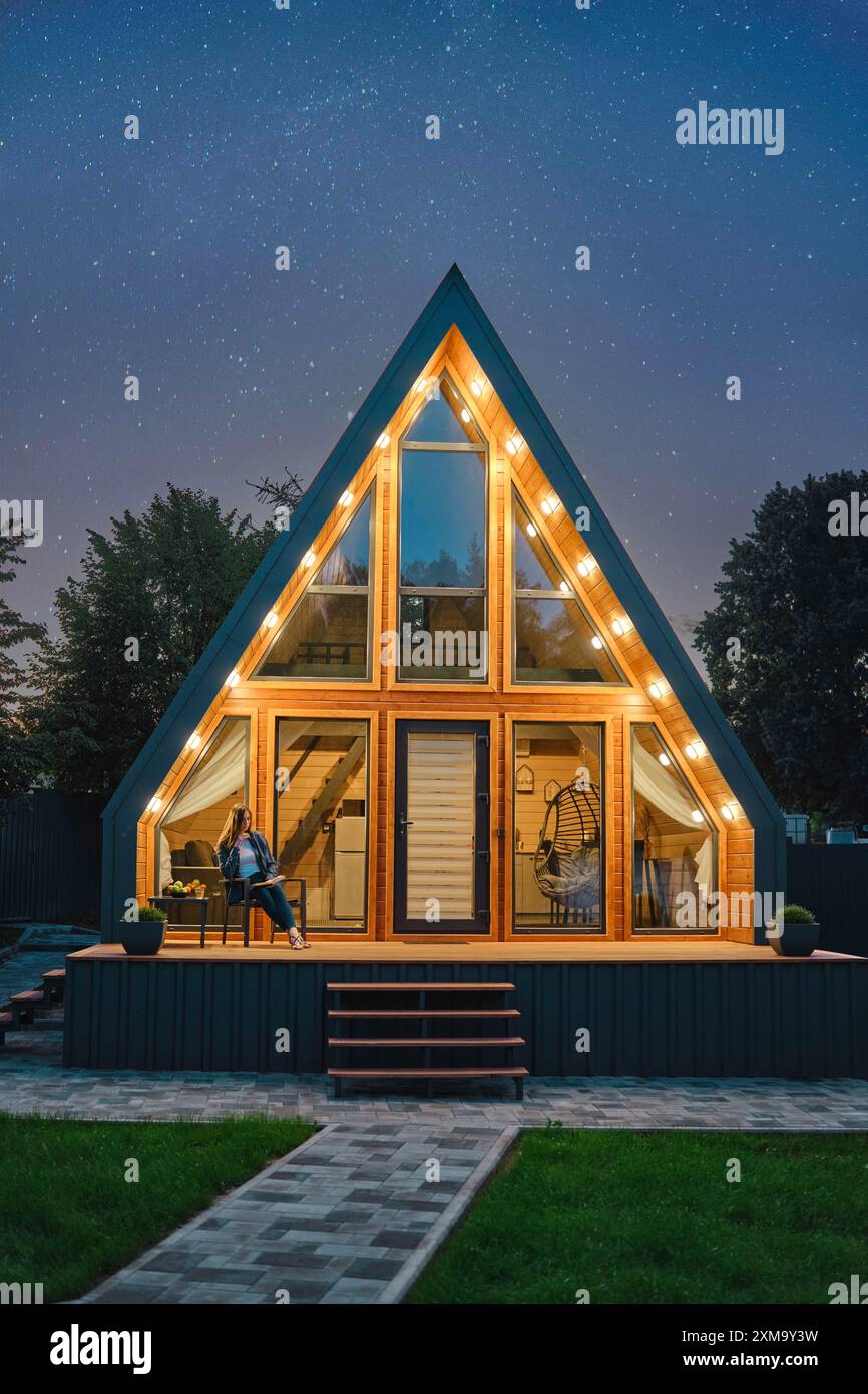 Une cabane en rondins triangulaire éclairée de l'intérieur se dresse sous un ciel étoilé. La lueur chaude de l'éclairage intérieur contraste avec le bleu frais de Banque D'Images