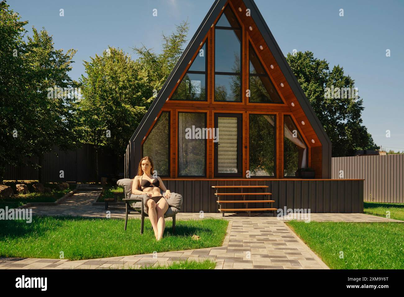 Une femme profitant de la tranquillité d'une journée ensoleillée, entourée d'une végétation luxuriante et de la beauté architecturale d'une maison moderne à ossature en a avec de grandes fenêtres Banque D'Images Une femme profitant de la tranquillité d'une journée ensoleillée, entourée d'une végétation luxuriante et de la beauté architecturale d'une maison moderne à ossature en a avec de grandes fenêtres Banque D'Images