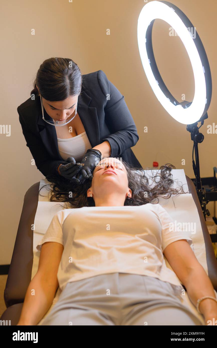 Un professionnel de la beauté effectuant un traitement sur une femme allongée sous une lumière annulaire, procédure de tatouage permanent des sourcils microblading, spa de beauté Banque D'Images