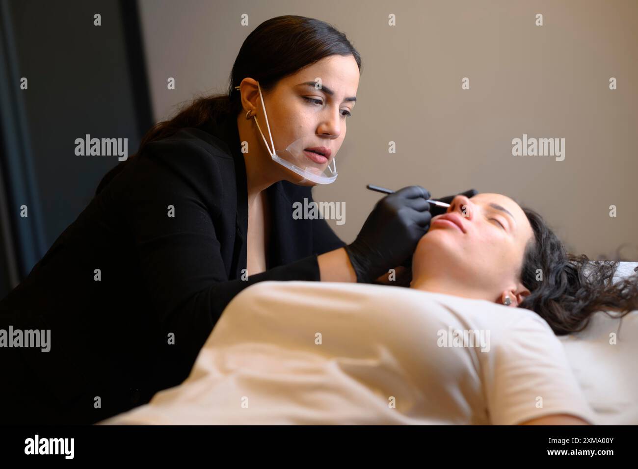 Cosmétologue en gants noirs et un masque facial effectuant un traitement des sourcils avec un crayon sur une femme couchée, microblading sourcil permanent Banque D'Images