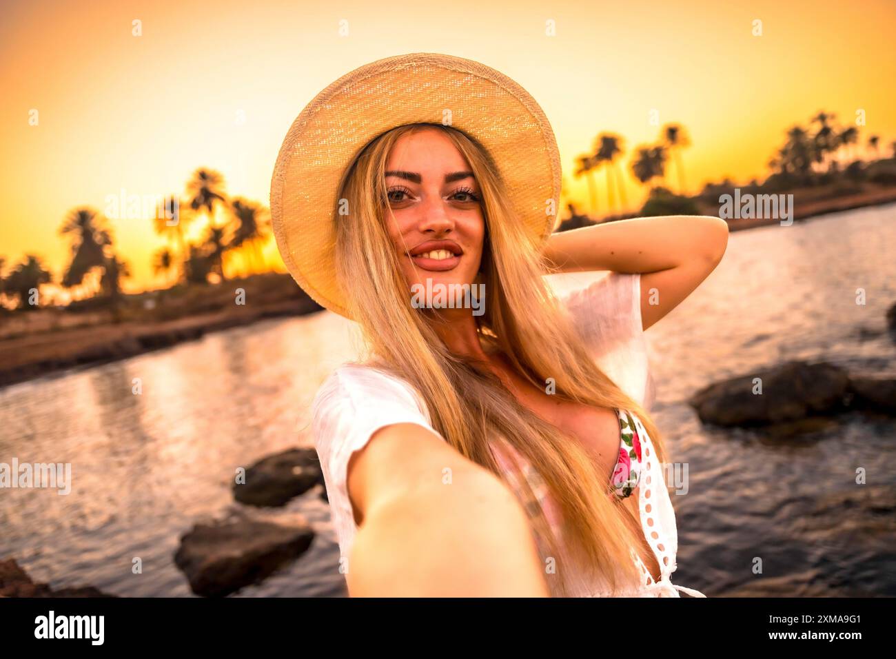 Point de vue personnel d'une femme blonde souriant à la caméra tout en prenant selfie pendant le coucher du soleil tropical Banque D'Images