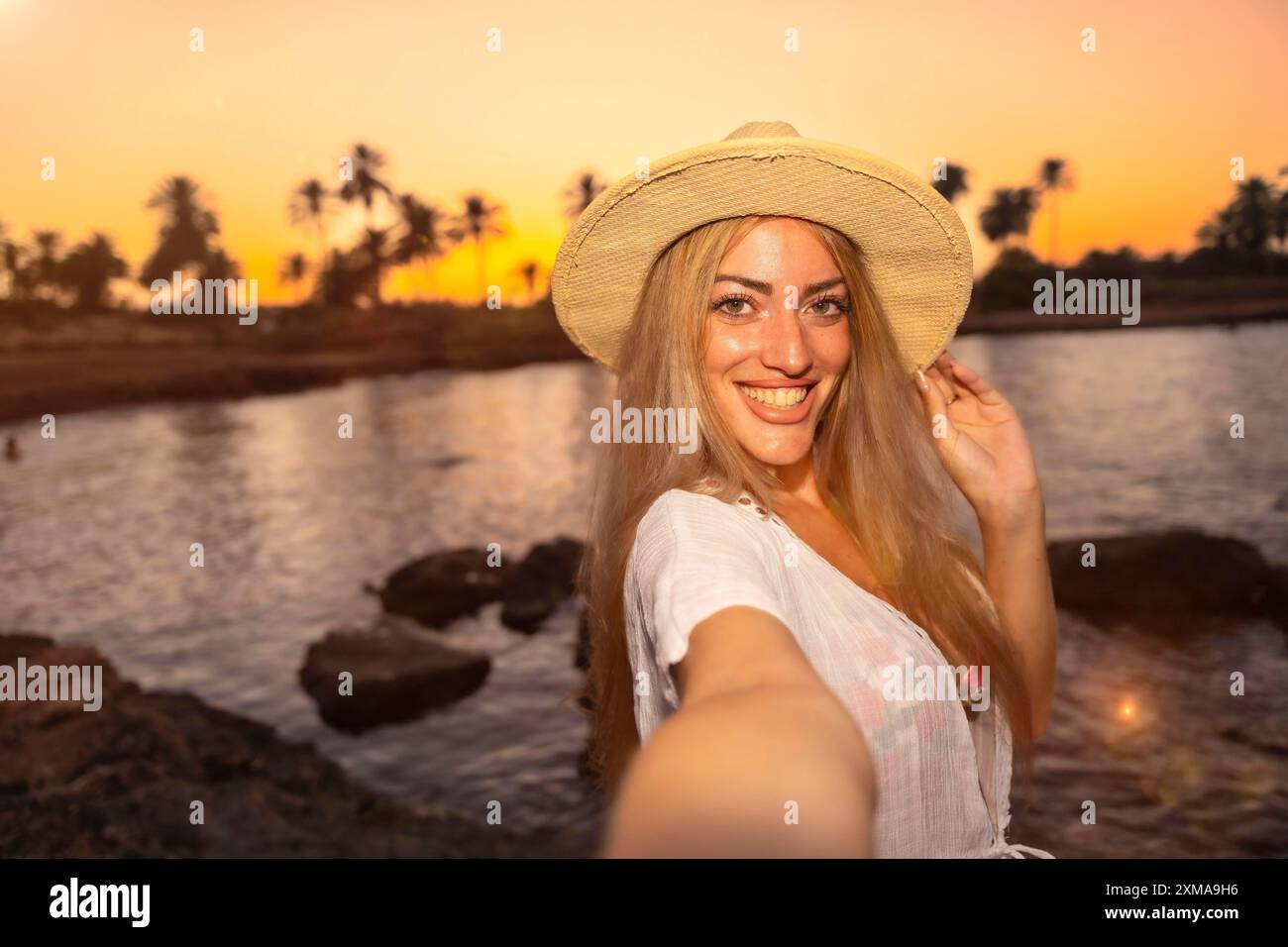 Point de vue personnel d'une femme avec chapeau de paille souriant tout en prenant selfie sur la plage pendant le coucher du soleil tropical Banque D'Images
