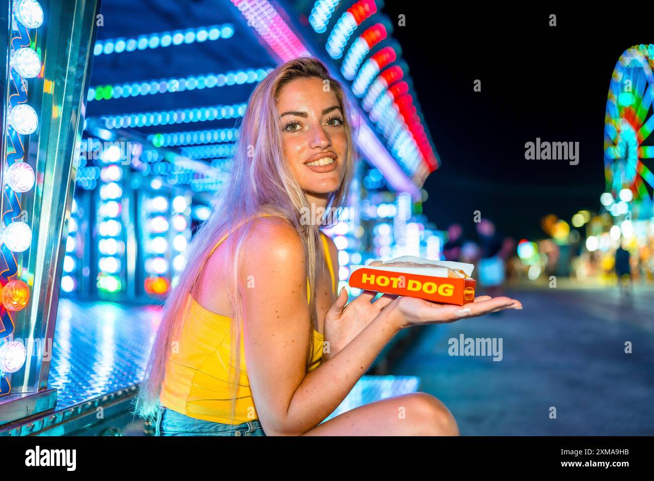 Femme maigre blonde mangeant un hot dog dans un parc d'attractions la nuit Banque D'Images