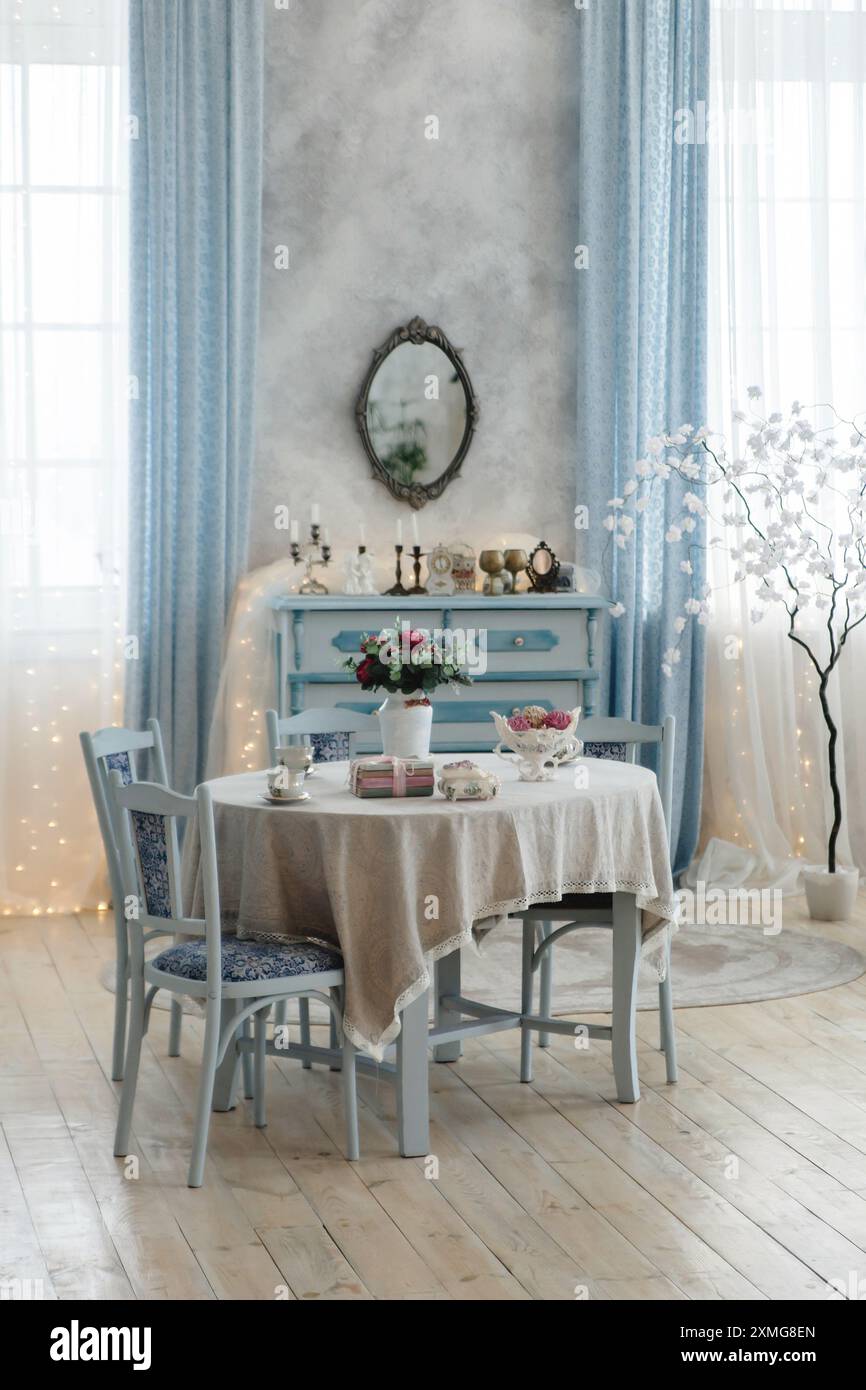 Salle à manger magnifiquement décorée avec une table vintage avec nappe en dentelle, des chaises bleues et une pièce maîtresse fleurie. Commode antique et miroir Banque D'Images