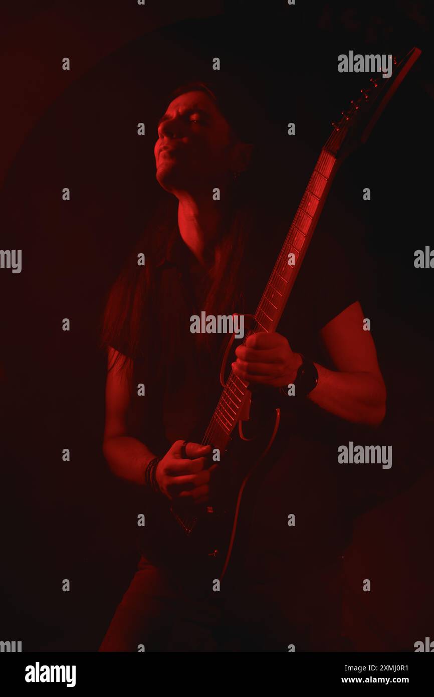 Portrait d'un guitariste métal aux cheveux longs jouant de la guitare électrique debout, illuminé par une lumière rouge pour un effet dramatique Banque D'Images