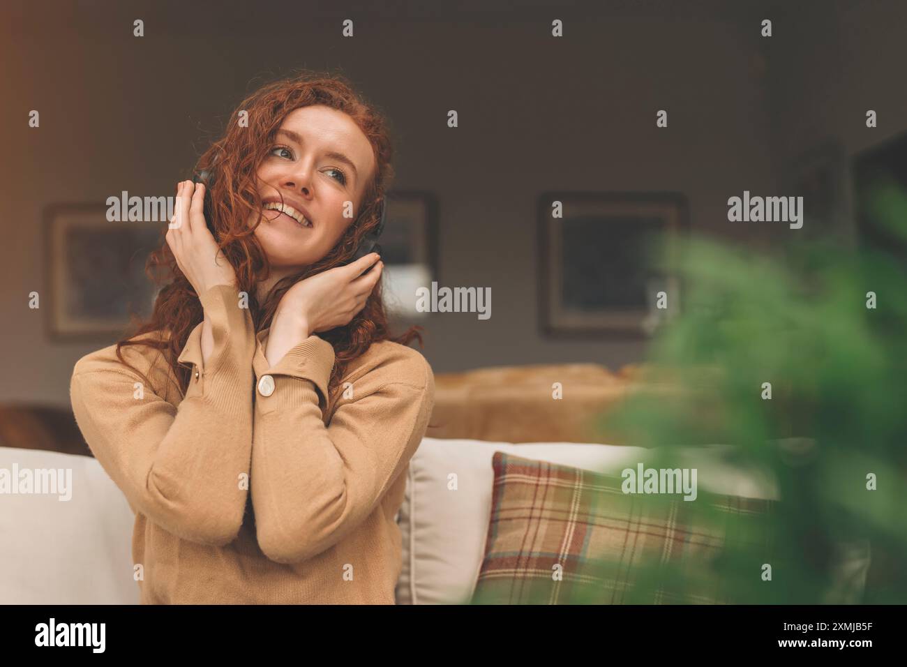 Heureuse jeune femme rousse en chemise brune écoutant des chansons préférées, des livres audio, des podcasts sur des écouteurs tout en étant assis près d'un canapé à la maison Banque D'Images