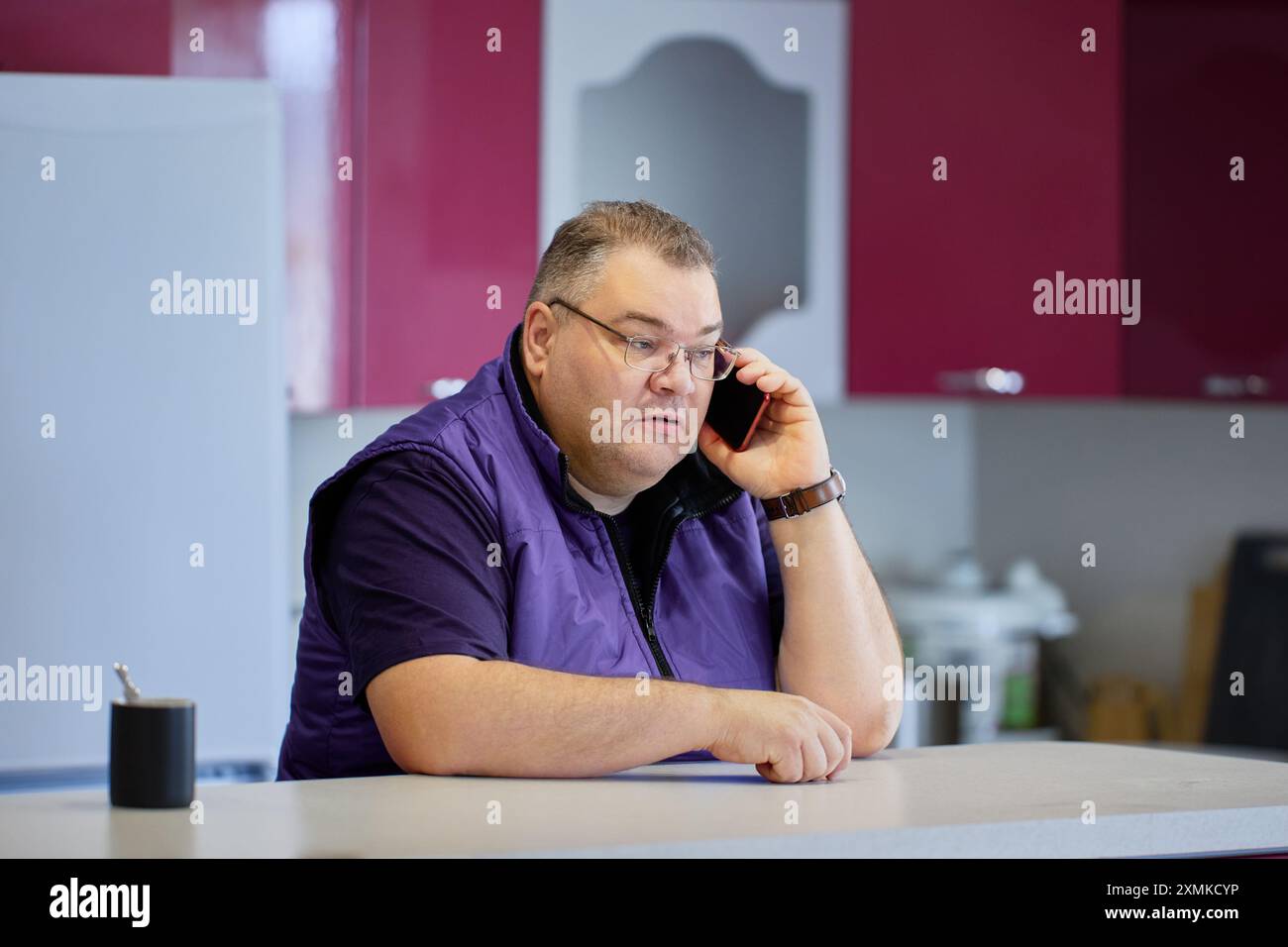 Conversation sérieuse au téléphone, grand homme caucasien dans ses années 40 avec des lunettes et les cheveux courts utilise smartphone dans la cuisine. Banque D'Images
