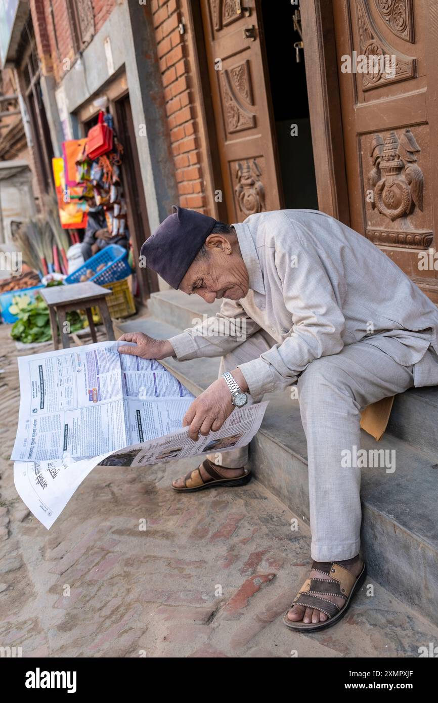 Un gentleman népalais portant une casquette bhadgaunle topi traditionnelle lit le journal dans les rues de Thimi, Katmandou, Népal. Banque D'Images