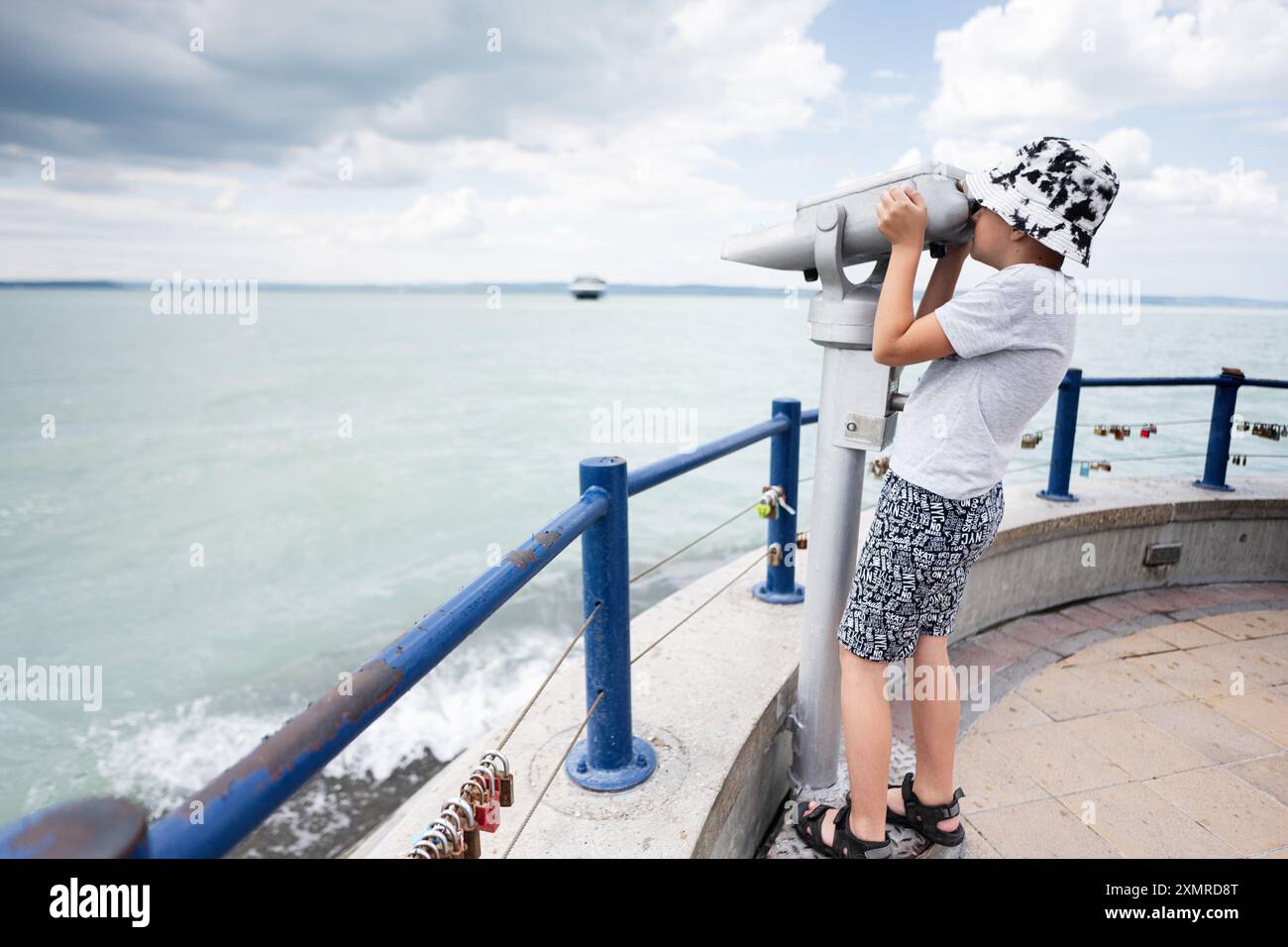 Enfant regardant à travers des jumelles à la mer, portant un chapeau, un jour nuageux. Vue sur la côte avec eau et balustrade. Banque D'Images