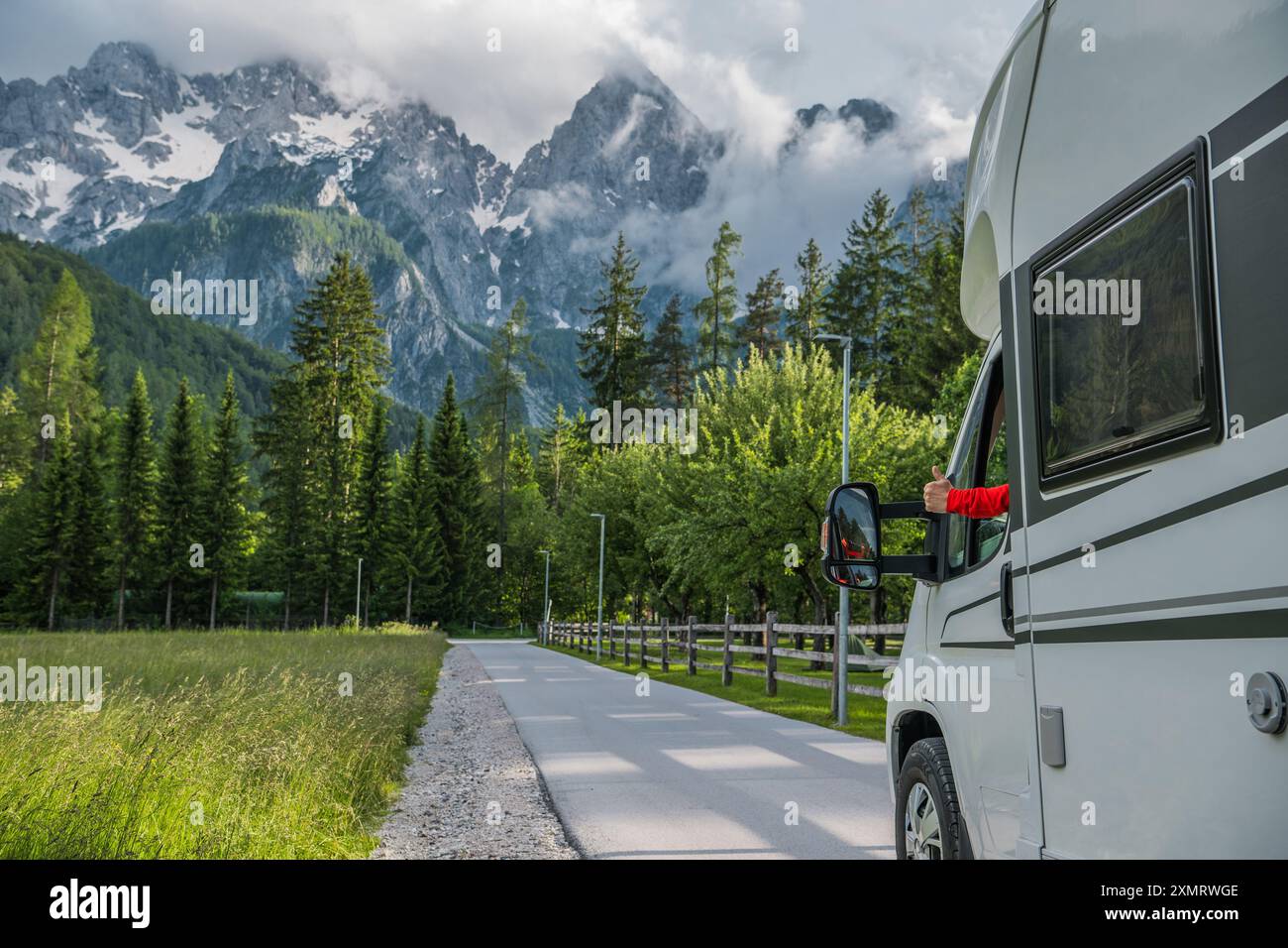 Un camping-car est garé le long d'une route sinueuse qui traverse une prairie verte vibrante, avec des montagnes majestueuses qui s'élèvent au-dessus. Le ciel nuageux ajoute Banque D'Images