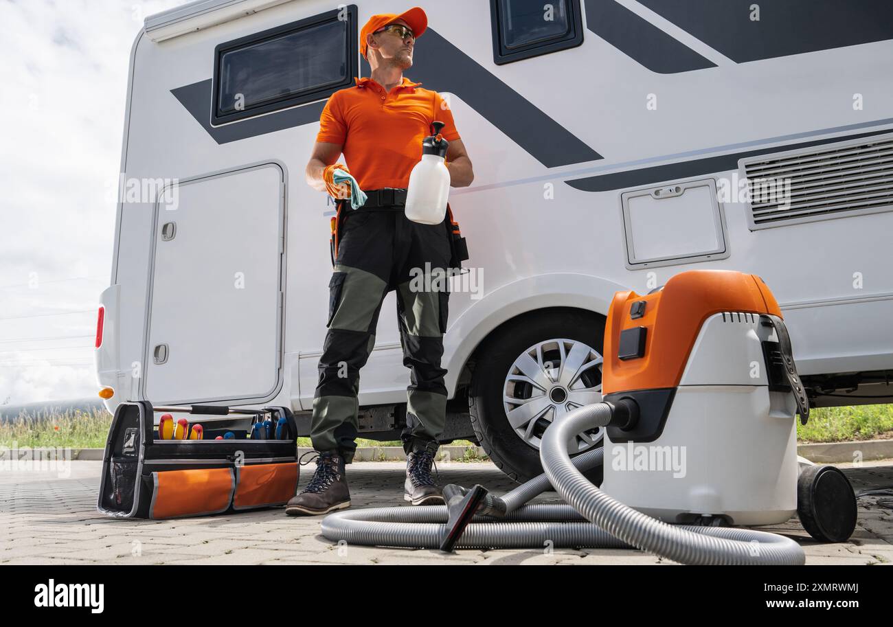 Un travailleur en uniforme orange se tient près d'un camping-car, tenant des fournitures de nettoyage. Il est entouré d'outils de nettoyage et d'équipement, prêt à l'entretien th Banque D'Images