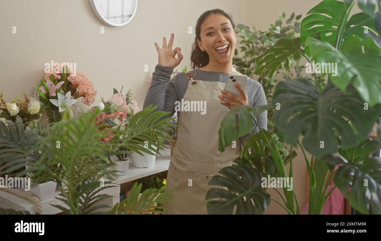 Femme hispanique faisant signe OK tout en tenant un smartphone dans un magasin de fleurs luxuriant avec des plantes vertes Banque D'Images