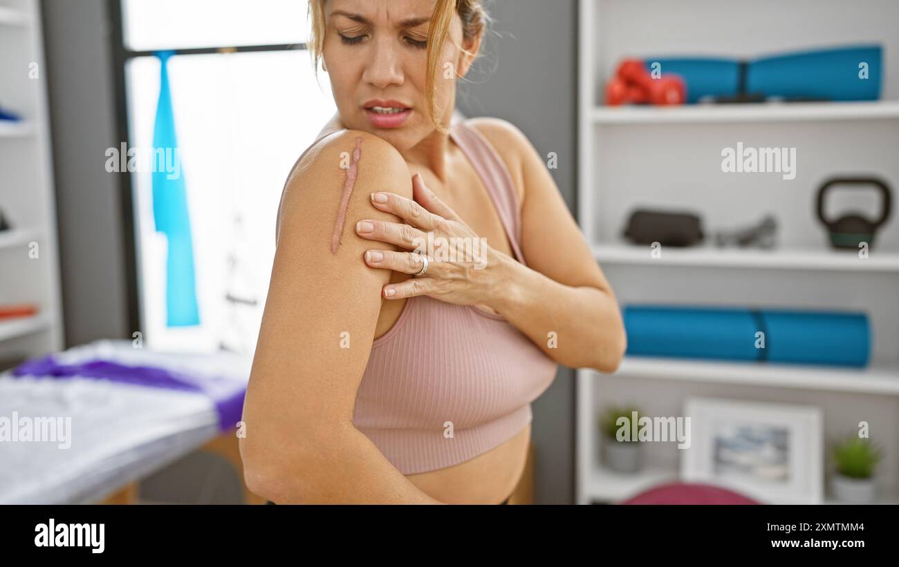 Une femme hispanique examine une cicatrice sur l'épaule dans une salle de clinique de réadaptation, décrivant la récupération et les soins de santé. Banque D'Images