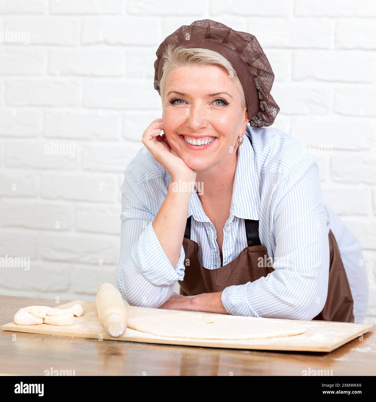 belle et heureuse femme dans le tablier et le chapeau de cuisinier souriant joyeux à la cuisine à la maison avec rouleau à pâtisserie et pâte sur la table regardant la caméra Banque D'Images