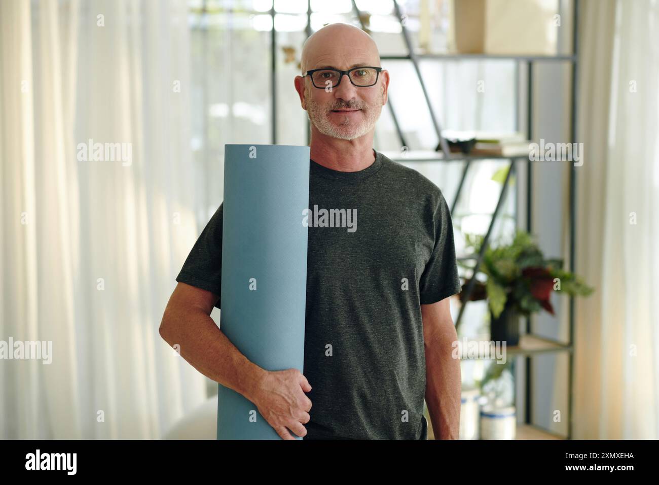 Portrait d'un homme caucasien âgé souriant tenant un tapis de yoga tout en se tenant debout dans la chambre avec des plantes et des étagères, portant des lunettes et des vêtements décontractés Banque D'Images