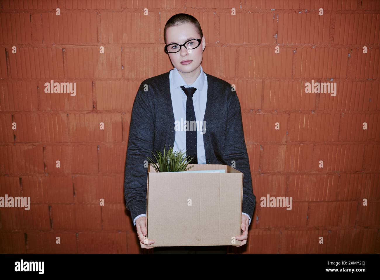 Employé féminin confiant avec buzz coupe de cheveux tenant boîte de trucs posant pour la caméra contre le mur de briques, espace de copie Banque D'Images