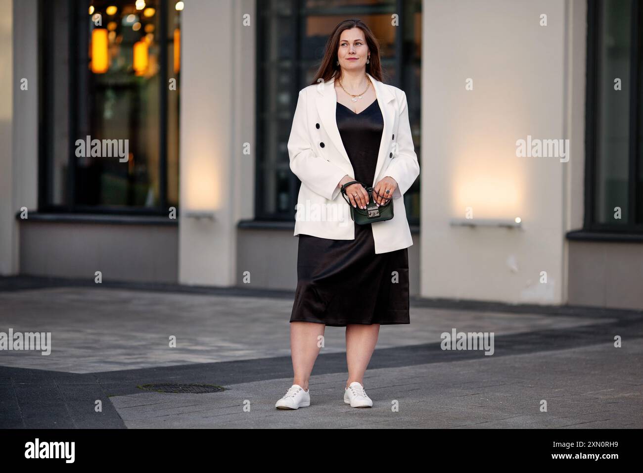 Jeune femme de taille plus se tient à l'extérieur dans un cadre urbain à la ville, portant une robe noire avec blazer blanc double-croisé, tenue avec des baskets blanches et Banque D'Images