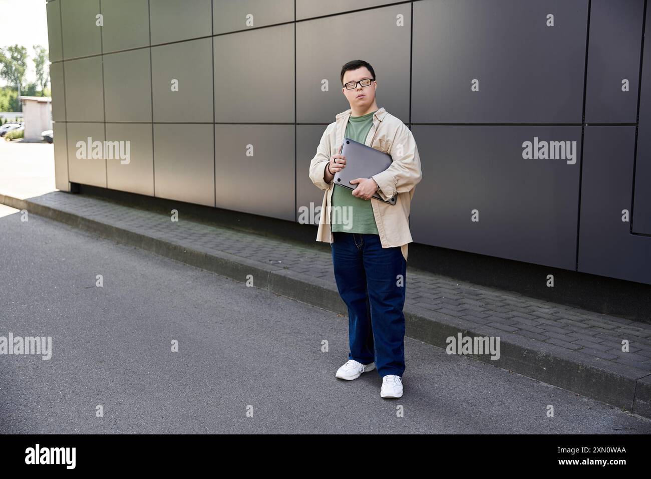 Un jeune homme atteint du syndrome de Down se tient devant un bâtiment moderne, tenant un ordinateur portable. Banque D'Images