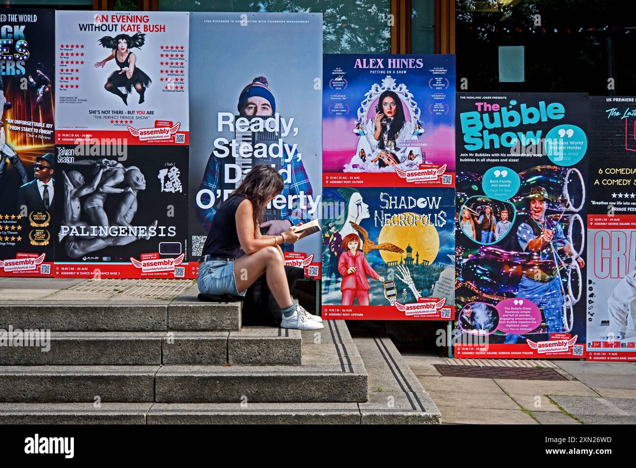 Jeune femme assise sur les marches lisant avec des affiches Edinburgh Fringe derrière elle. Banque D'Images