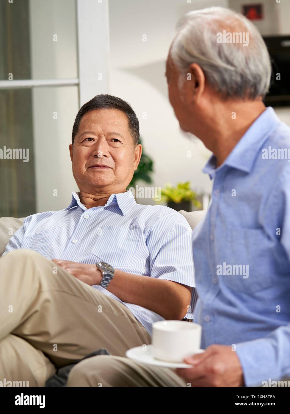 deux heureux hommes asiatiques seniors assis et discutant sur le canapé de la famille à la maison avec des épouses en arrière-plan Banque D'Images