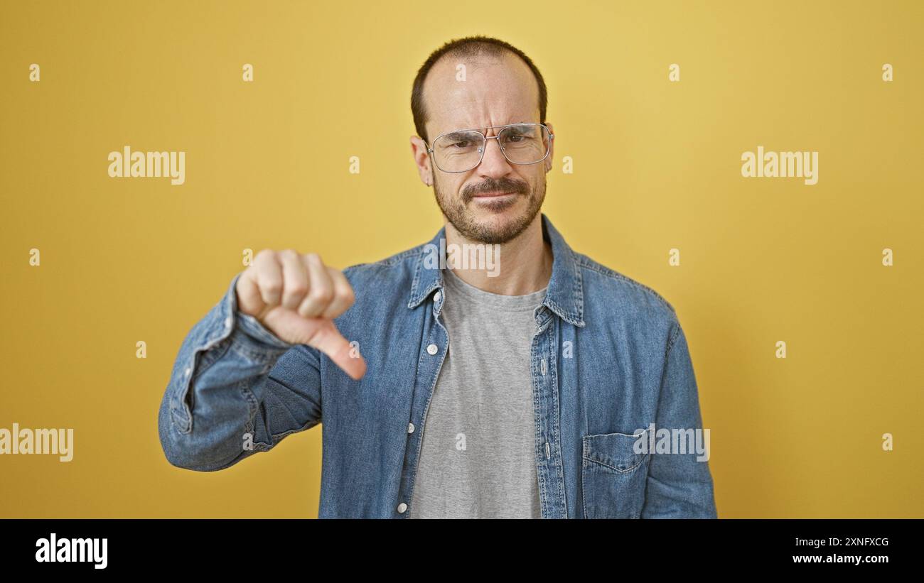 Un homme barbu et chauve portant des lunettes et une chemise en Jean exprimant sa désapprobation avec un geste de pouce vers le bas sur un fond jaune. Banque D'Images