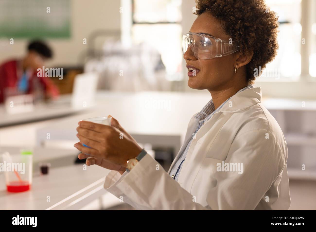 Au lycée, enseignant portant une blouse de laboratoire et des lunettes de sécurité tenant un bécher en classe Banque D'Images