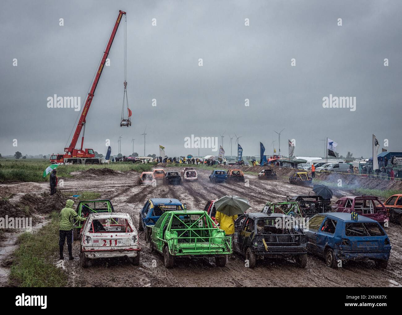 Course de voitures de démolition dans des conditions boueuses. Les spectateurs avec des parapluies regardent une grue soulever une voiture au milieu de l'action. Banque D'Images