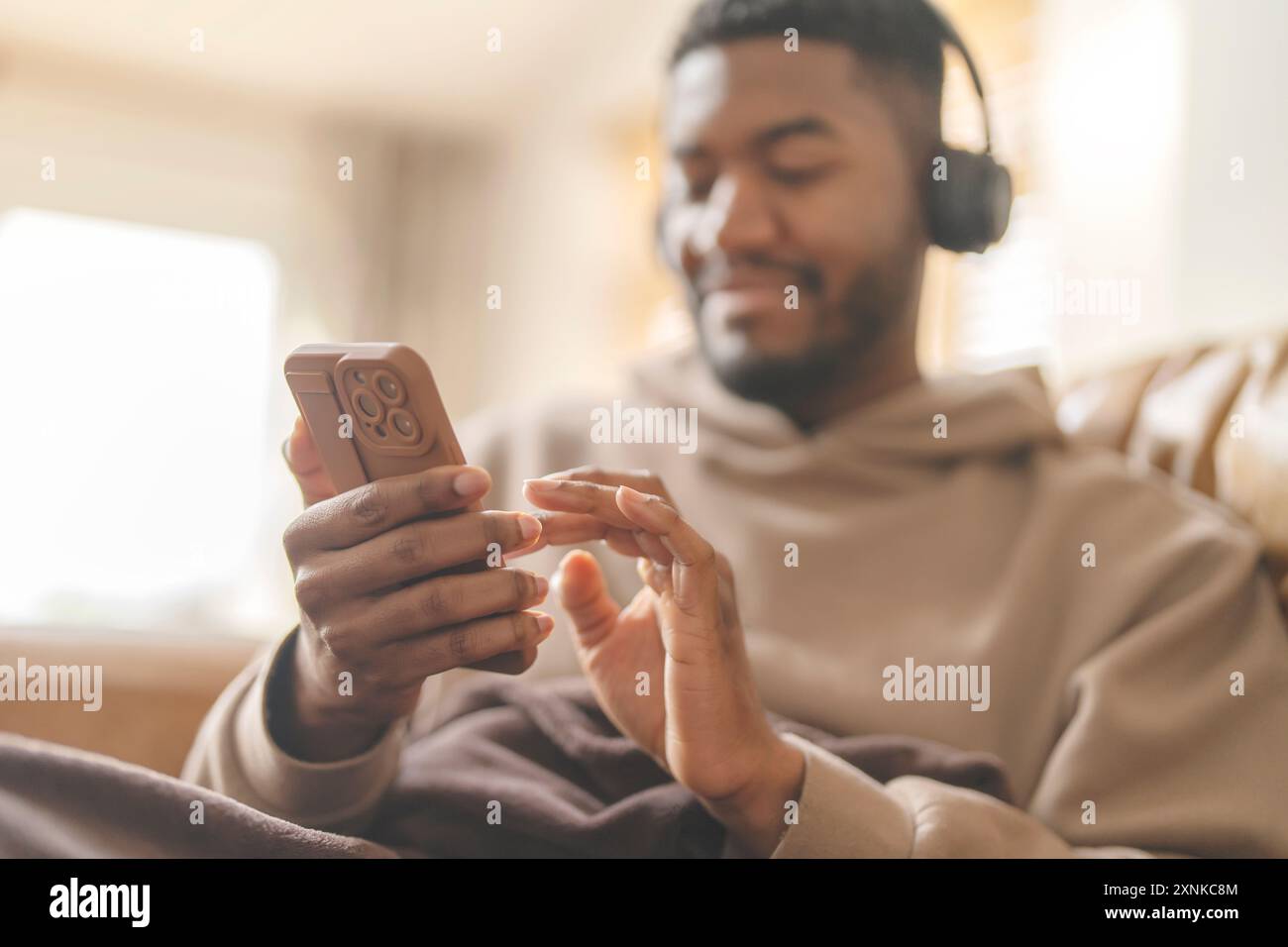 Homme heureux dans le sweat à capuche brun en utilisant le téléphone, étudier, apprendre, faire des achats, travailler en ligne, parler, écouter de la musique, livre audio, podcast tout en étant assis Banque D'Images