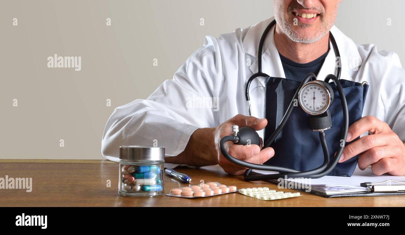 Détail d'un médecin spécialiste souriant montrant un tensiomètre sur une table de consultation avec des médicaments et un fond avec fenêtre avec des plantes. Banque D'Images