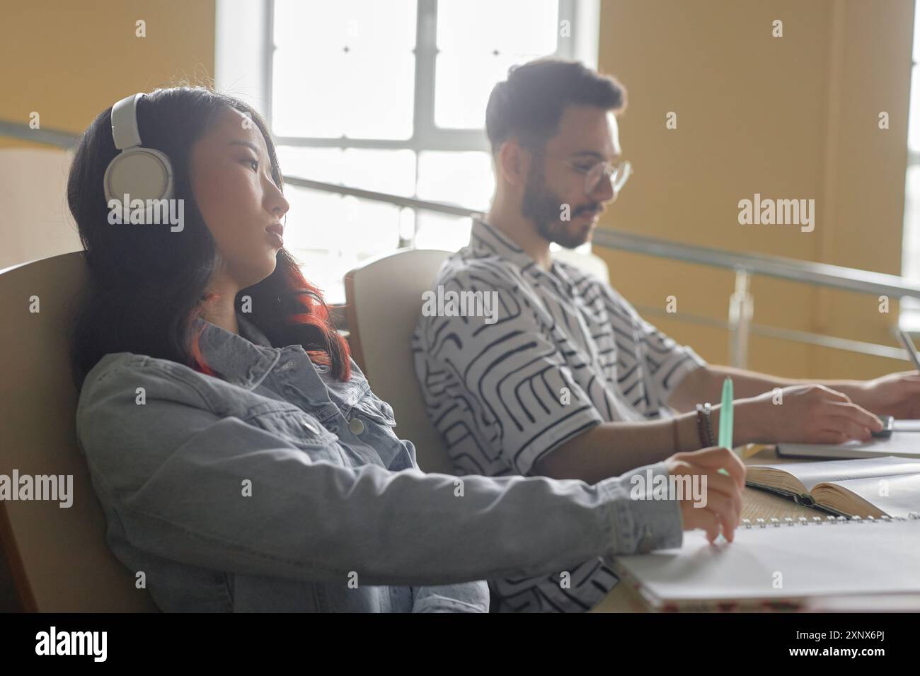 Vue latérale de l'étudiante asiatique fatiguée dans des écouteurs au bureau à côté de la fenêtre assistant à une conférence ou écrivant un essai avec de la musique à l'université Banque D'Images
