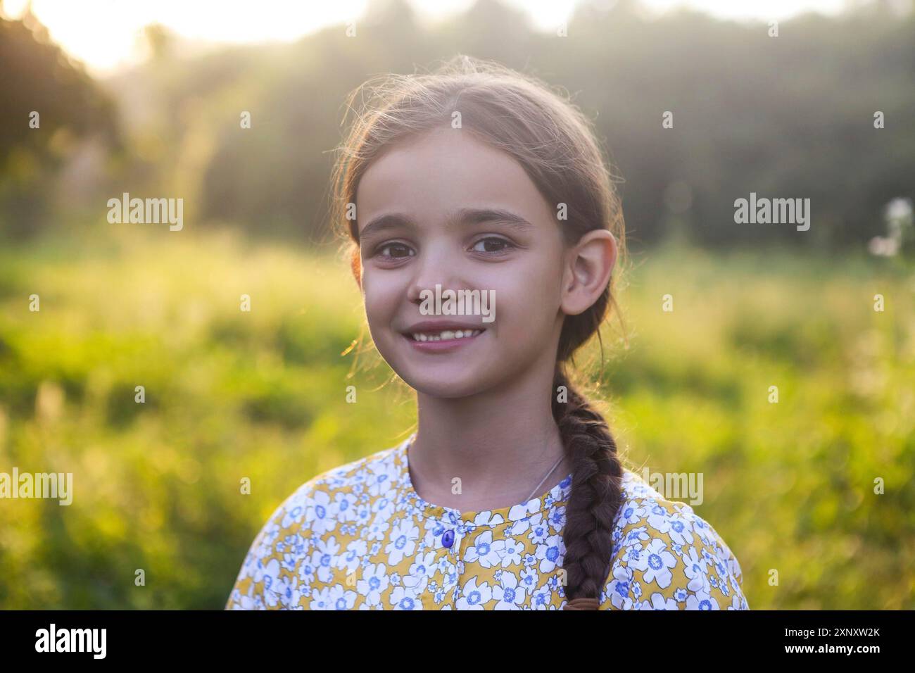 Petite fille gaie avec queue de porc dans des vêtements colorés souriant et regardant l'appareil photo tout en se tenant dans le champ vert en été soir Banque D'Images