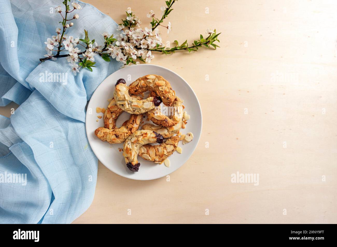 Biscuits en croissant d'amande sur une assiette, branche de fleur de ressort et serviette bleue sur une table en bois clair, grand espace de copie, vue d'en haut Banque D'Images