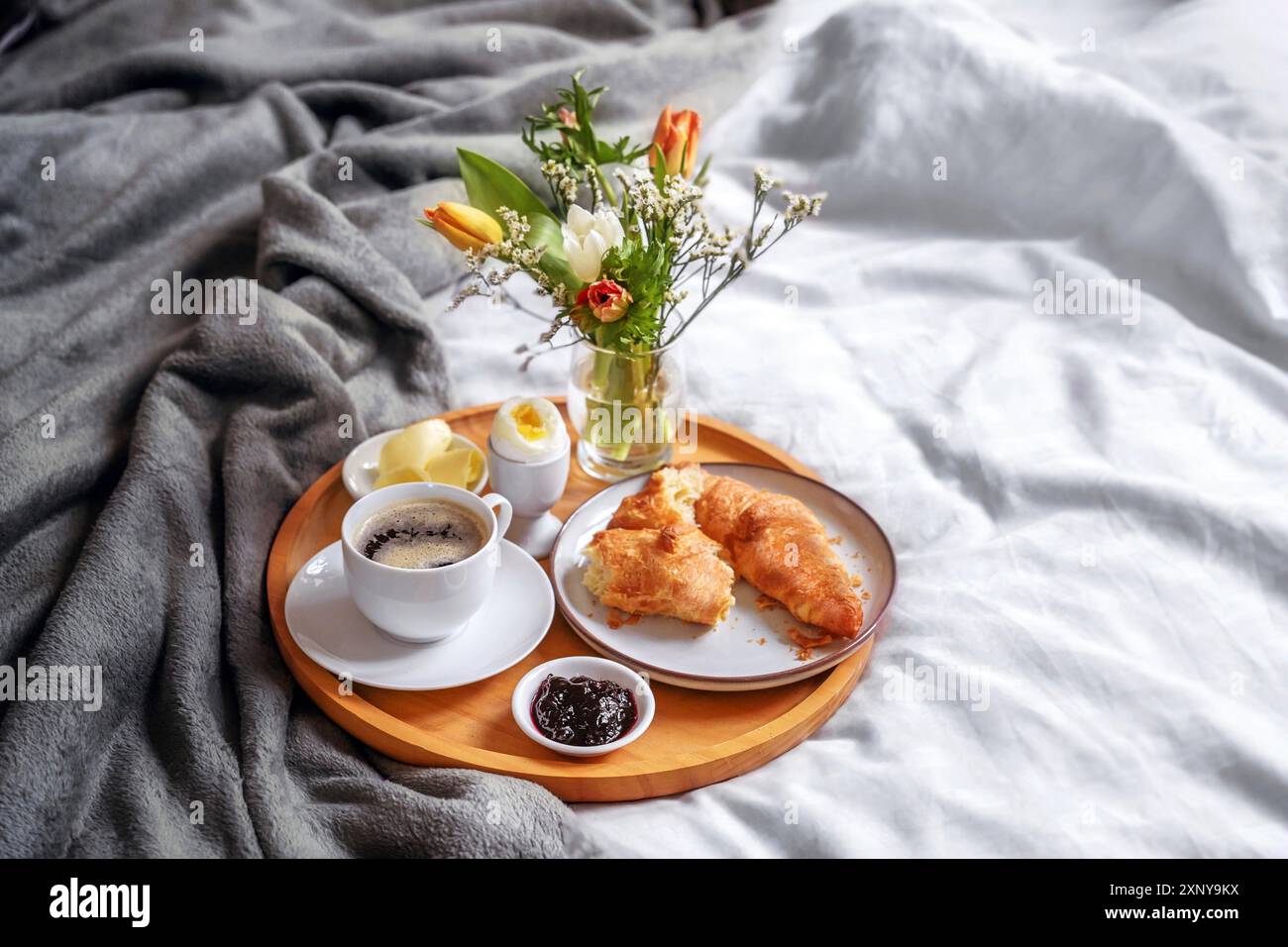 Plateau petit déjeuner en bois avec croissant, café, confiture, œuf et bouquet de fleurs fraîches servi sur le lit avec linge de lit clair et couverture grise, vacances ou week-end Banque D'Images Plateau petit déjeuner en bois avec croissant, café, confiture, œuf et bouquet de fleurs fraîches servi sur le lit avec linge de lit clair et couverture grise, vacances ou week-end Banque D'Images