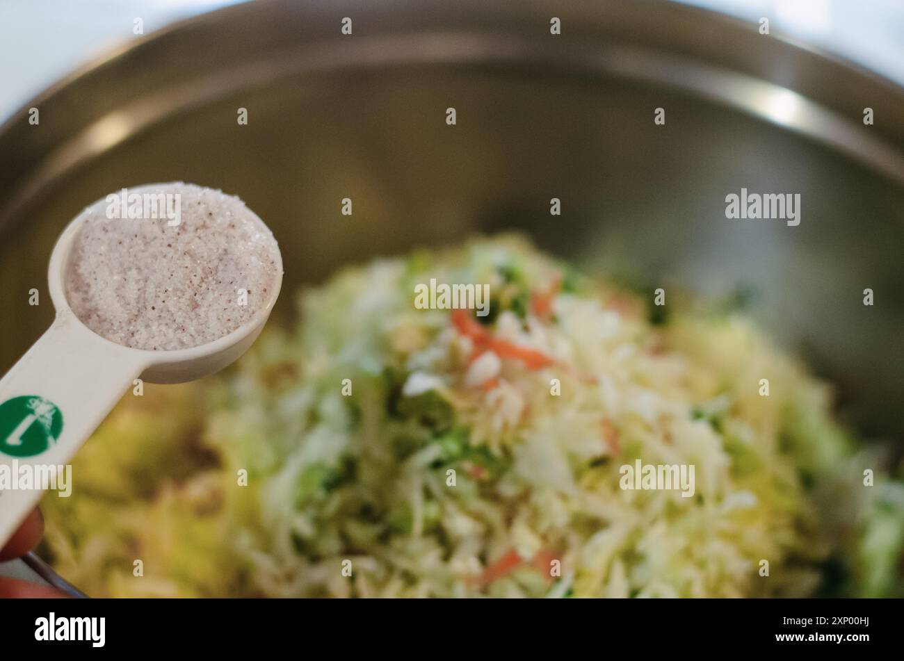 Une cuillère à mesurer pleine de sel est ajoutée à un mélange de choucroute dans un bol à mélanger en métal Banque D'Images