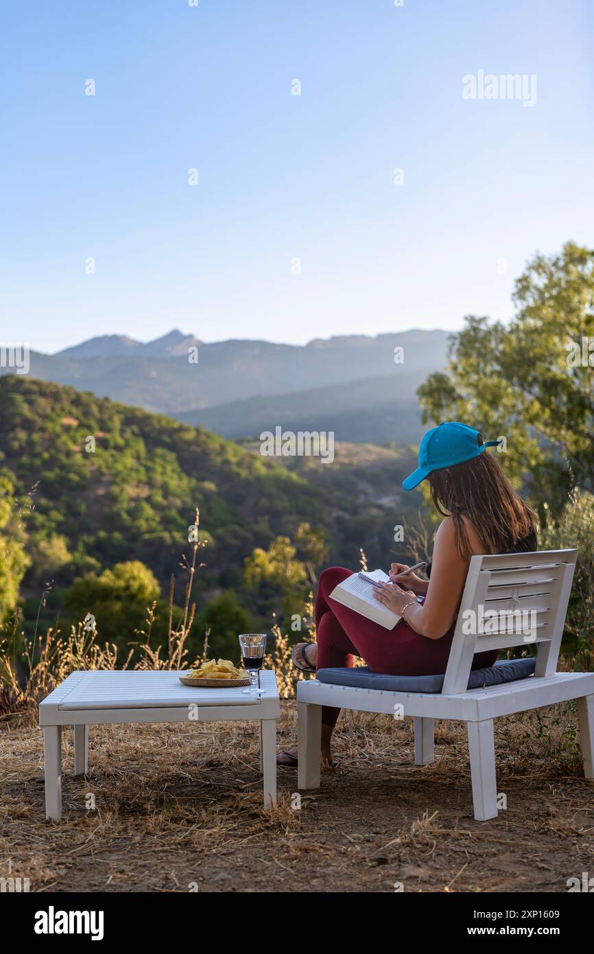 Jeune femme assise à l'extérieur, lisant un livre et profitant d'un paysage de montagne andalou au coucher du soleil. Sentiment de tranquillité et de bien-être chez une maman Banque D'Images