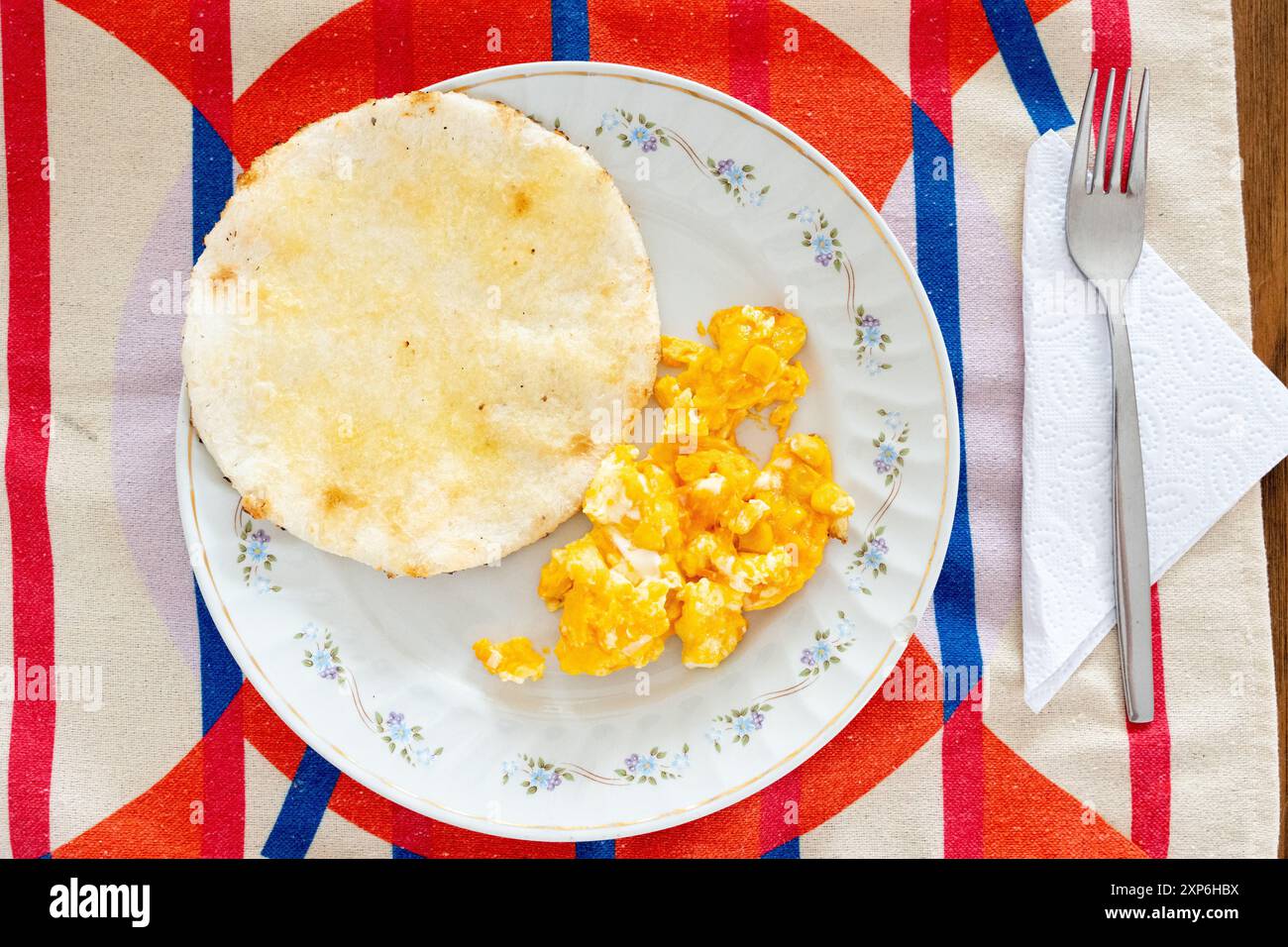 Une photo aérienne présentant un petit-déjeuner colombien typique, avec une assiette avec arepa et œufs brouillés sur une nappe colorée et vibrante Banque D'Images