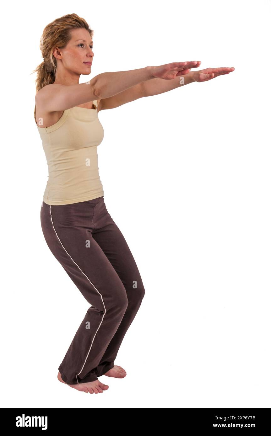 Vue de trois quarts de longueur du corps d'une jeune femme blonde en robe de sport montrant un squat d'exercice sportif avec les bras tendus isolés sur blanc. Banque D'Images