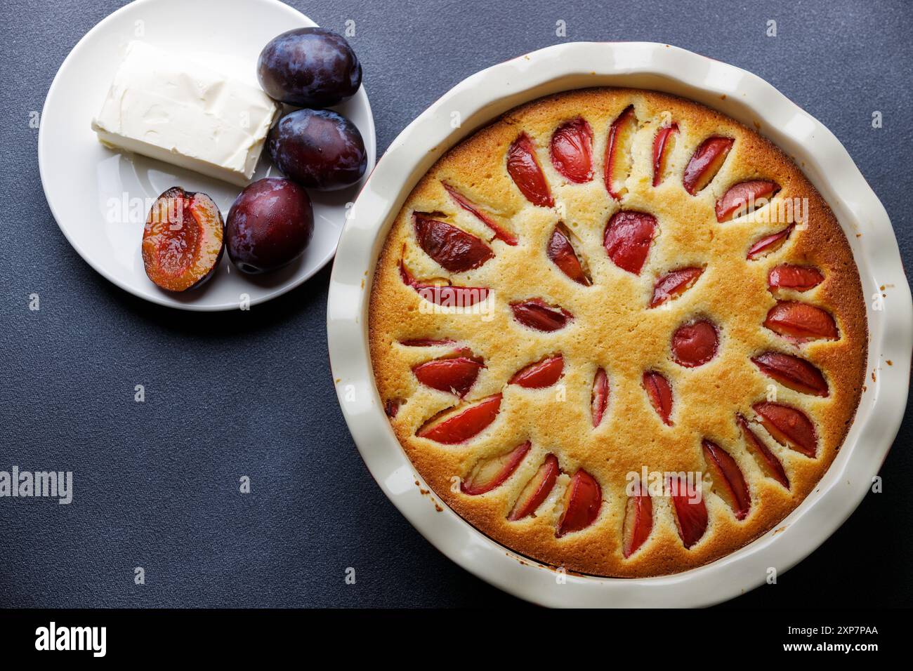 Un gâteau aux prunes magnifiquement cuit est servi avec un morceau de beurre et des prunes fraîches sur un comptoir sombre. Banque D'Images