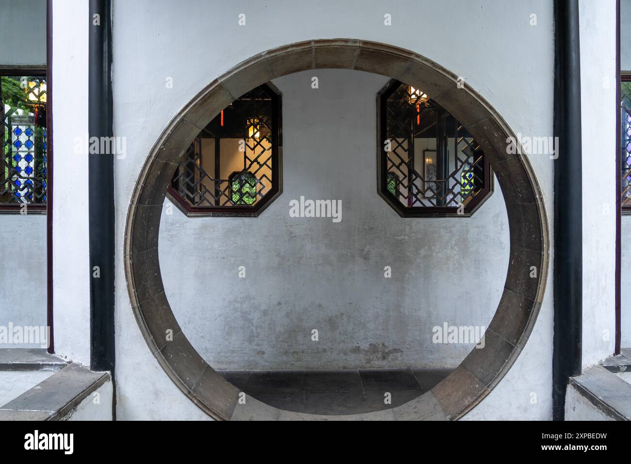 Suzhou, Chine - 11 juin 2024 : Une ouverture circulaire dans un mur blanc encadre deux fenêtres octogonales avec un treillis complexe. Banque D'Images