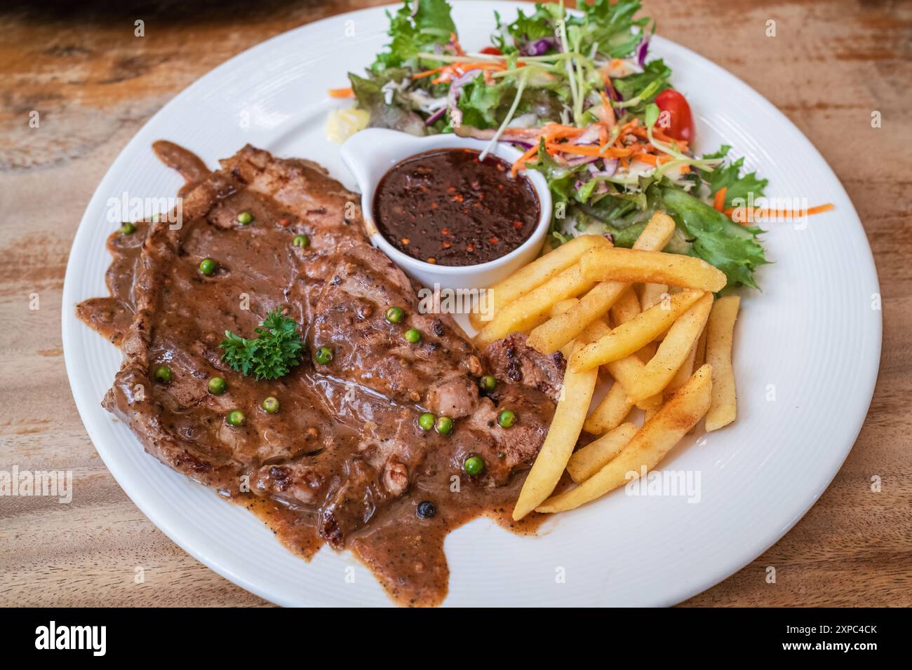 Steak de bœuf grillé avec frites, légumes et sauce piquante épicée dans une assiette blanche sur une table en bois Banque D'Images