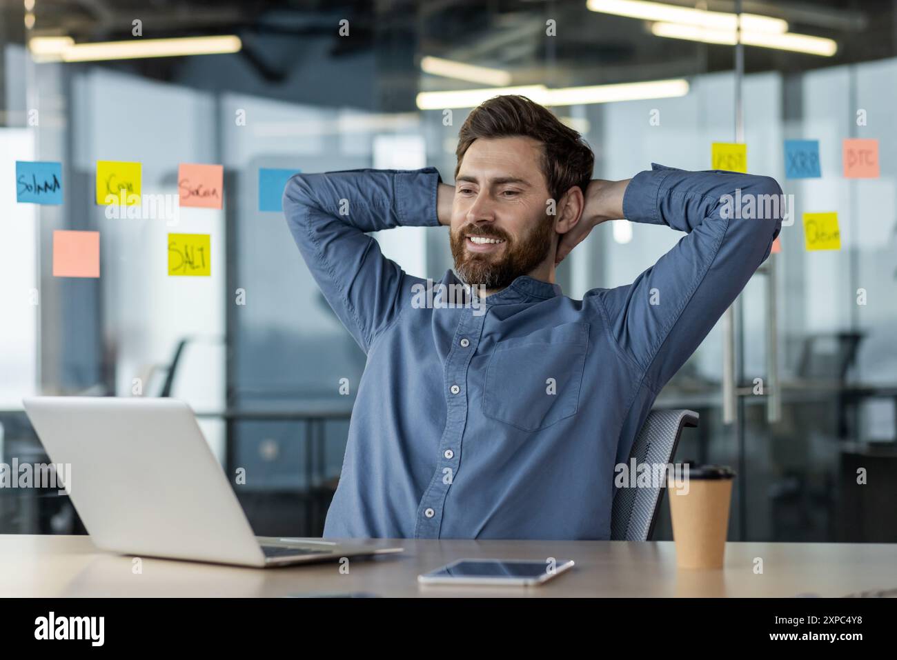 Homme d'affaires confiant prenant une pause détente au bureau. Homme assis avec les mains derrière la tête, souriant à l'ordinateur. Environnement de bureau avec des notes adhésives sur le mur de verre. Banque D'Images
